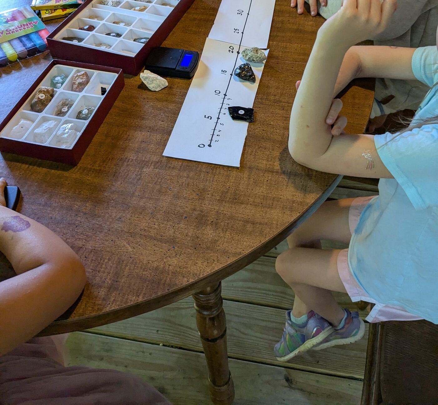 Trays of sorted rocks, a small digital scale and a paper number line on a table, arms of the children at work