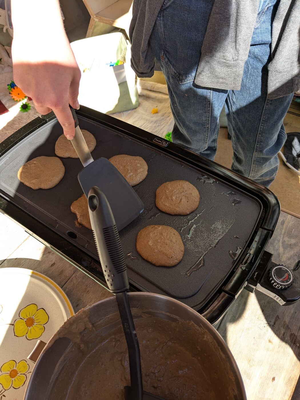 Pancakes cooking on an electric griddle, a spatula and a bowl of batter