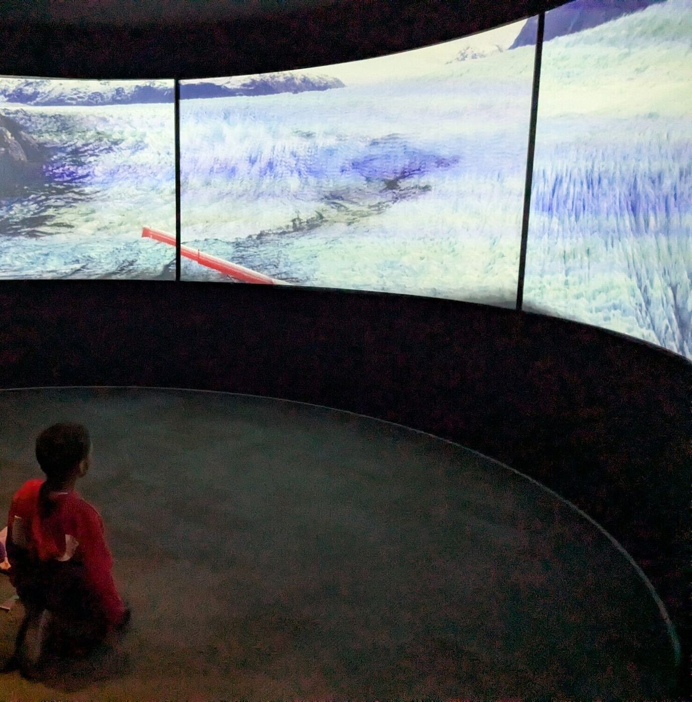 A child seen from behind, sitting before a wide projection screen at the science museum