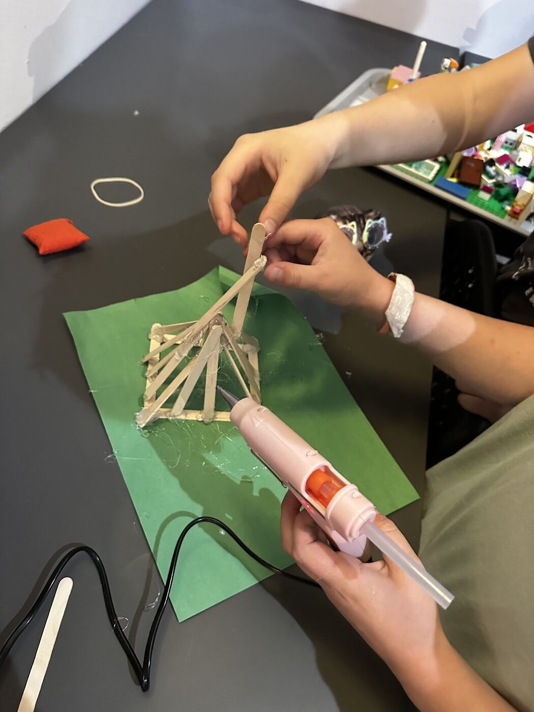 Hands holding a popsicle-stick tower steady while another hand works the glue gun