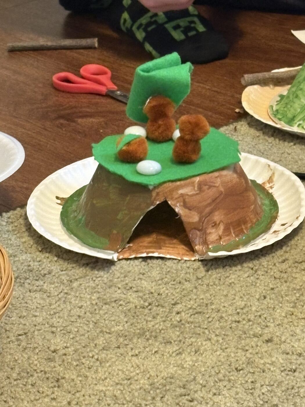 A paper-plate den with a felt roof and pom-pom animals