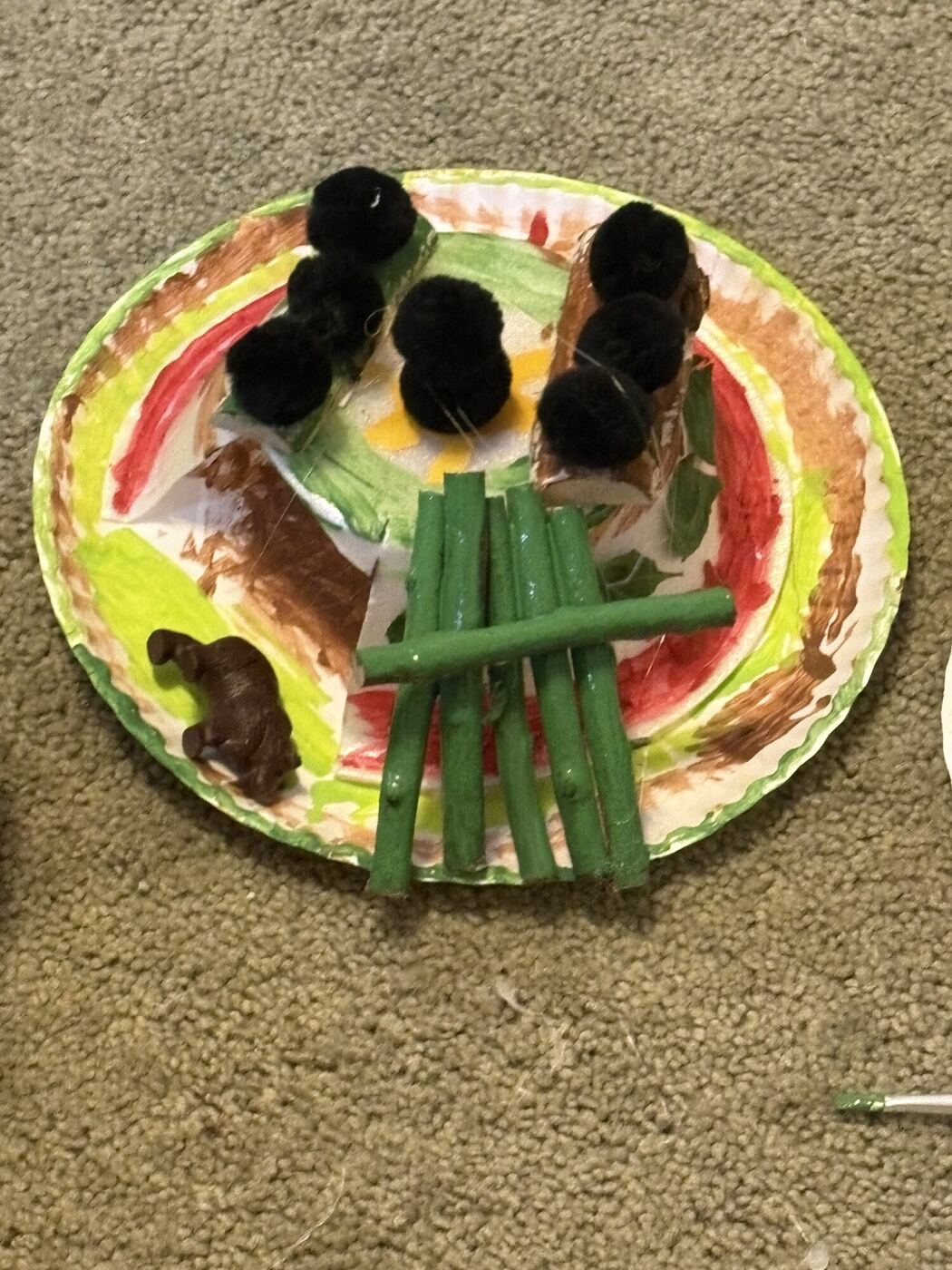 A painted paper-plate den with black pom-poms and green sticks