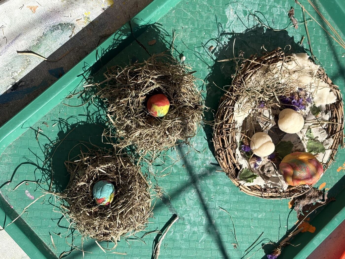 Three nests of dry grass on a green tray, each with painted eggs