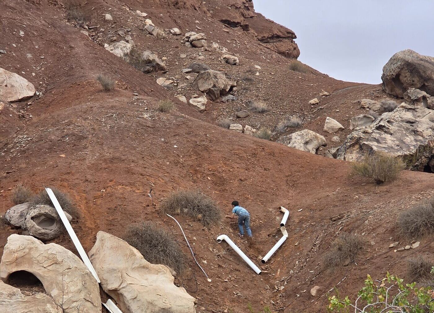 White boards laid out on a red dirt slope among boulders, one child seen from behind climbing up to them