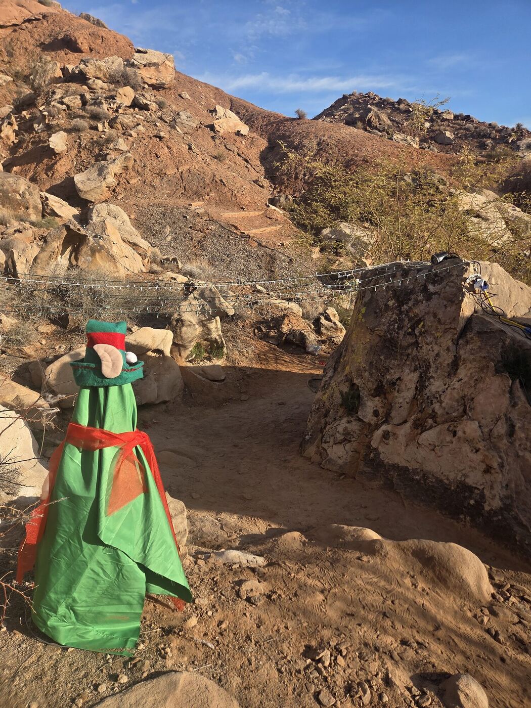 A cloth elf standing beside a desert path among boulders, a string of lights behind it