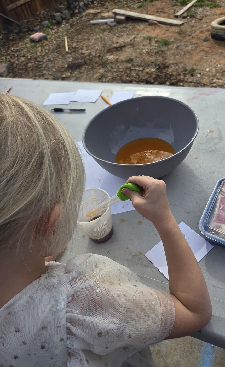 A child seen from behind squeezing a dropper over a cup, an orange soda in a bowl beyond