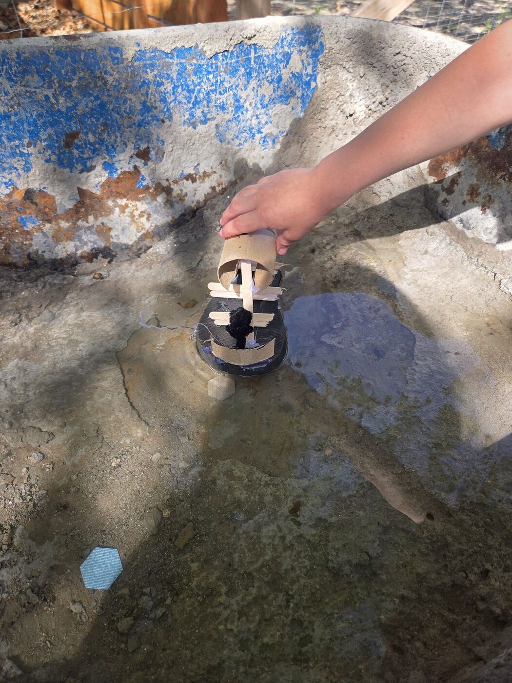 A hand setting a small boat of cardboard tubes and sticks into water in an old basin