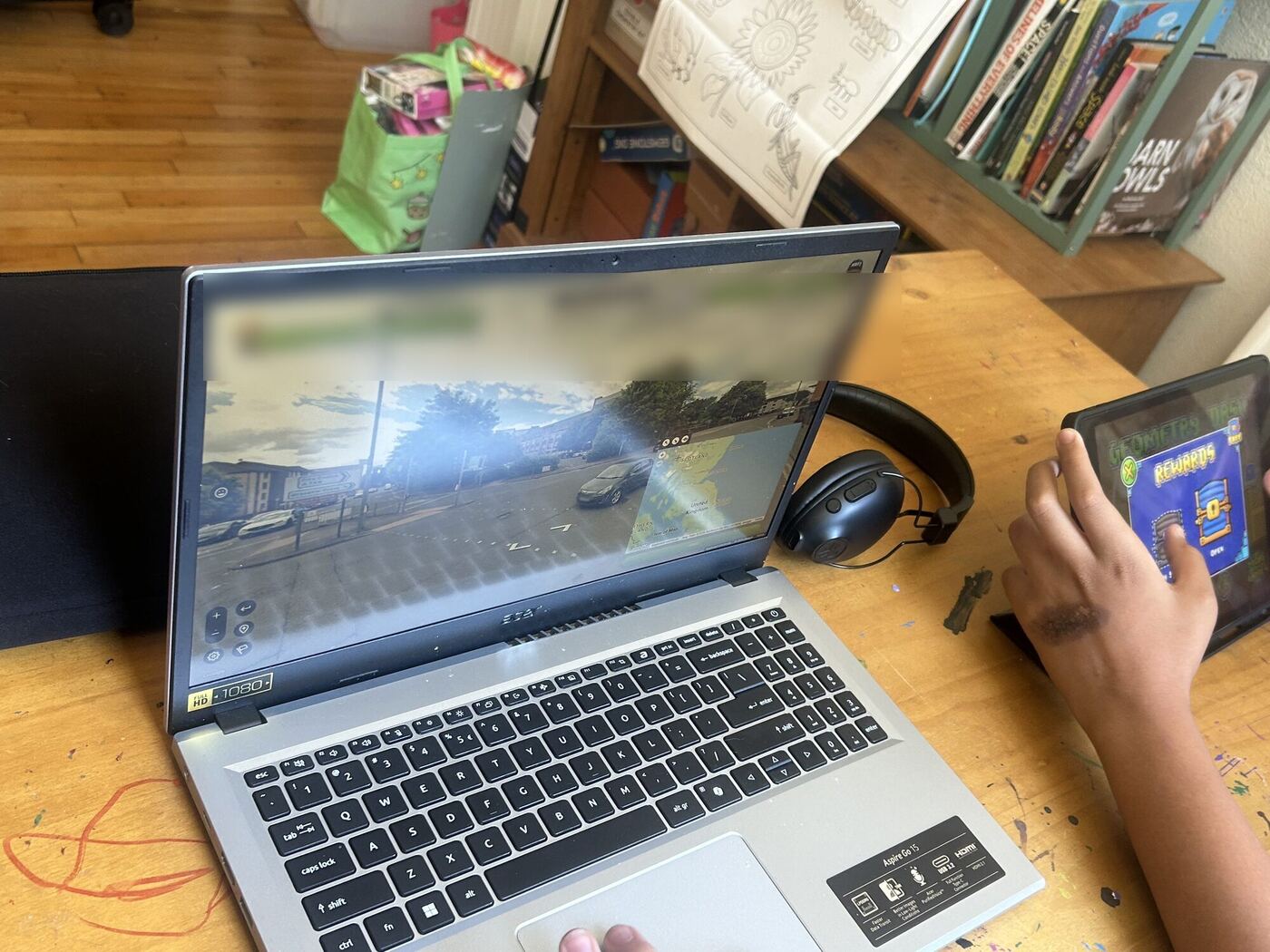 A laptop showing a street-view geography game, a hand on a tablet beside it