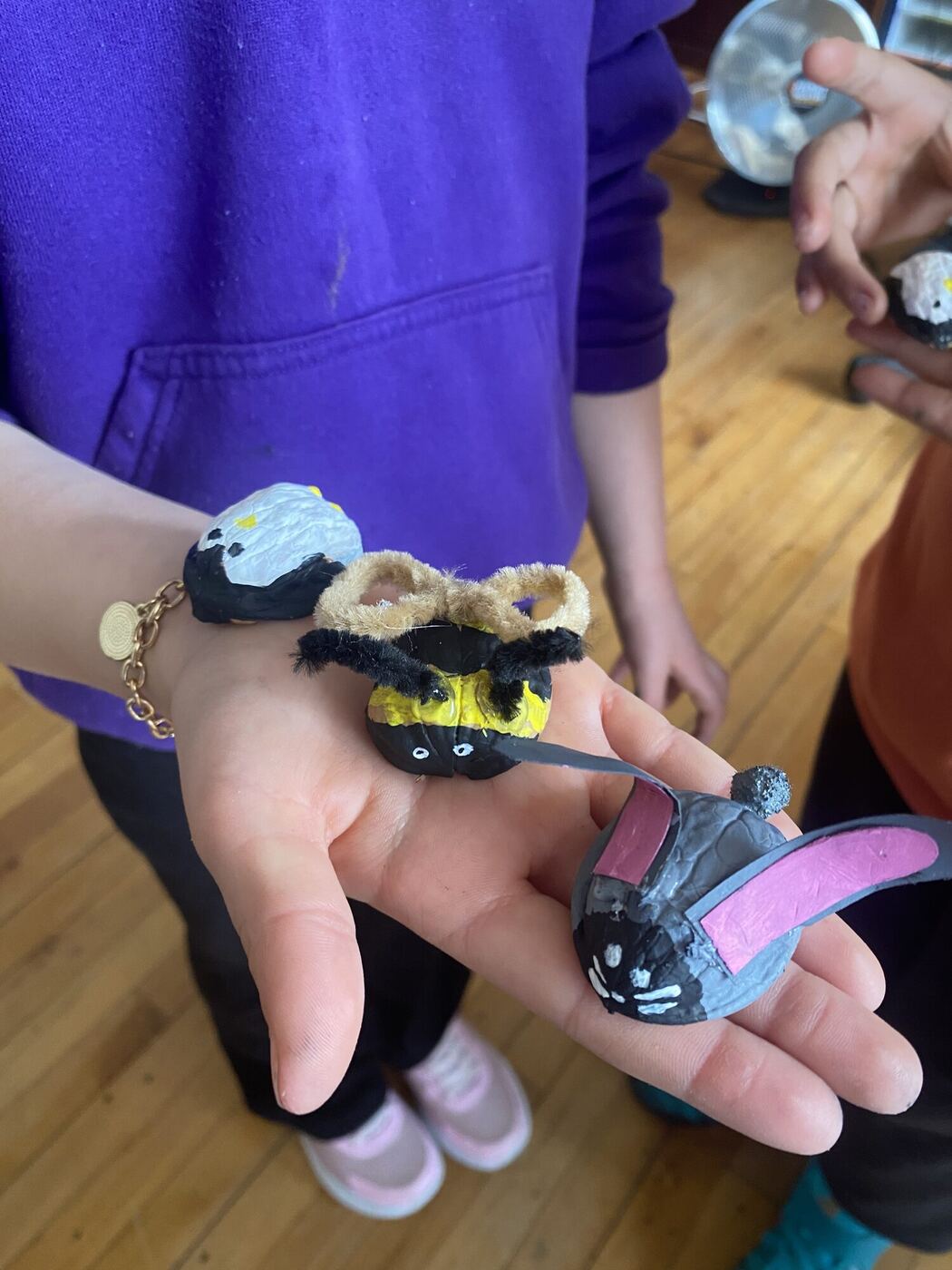 An open hand holding three painted walnut-shell creatures: a bee, a bunny and a bird