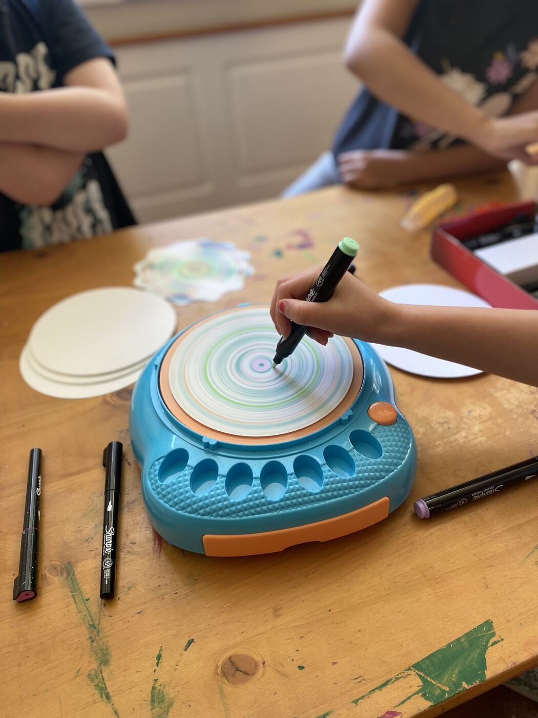 A hand holding a marker to a paper disc on a spinning art machine, finished discs beside it