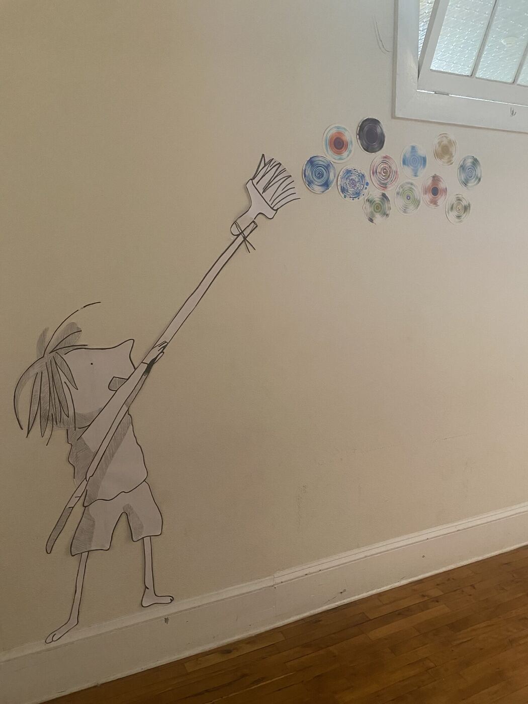 A drawn figure on a hallway wall reaching a brush up to a cluster of colored dot paintings