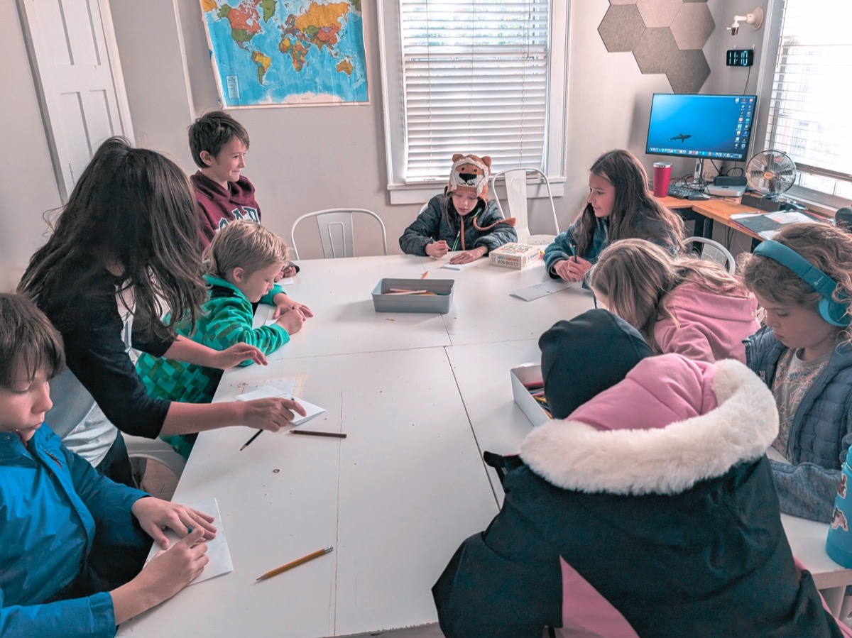 A group of learners writing around one long shared table, an adult leaning in beside them