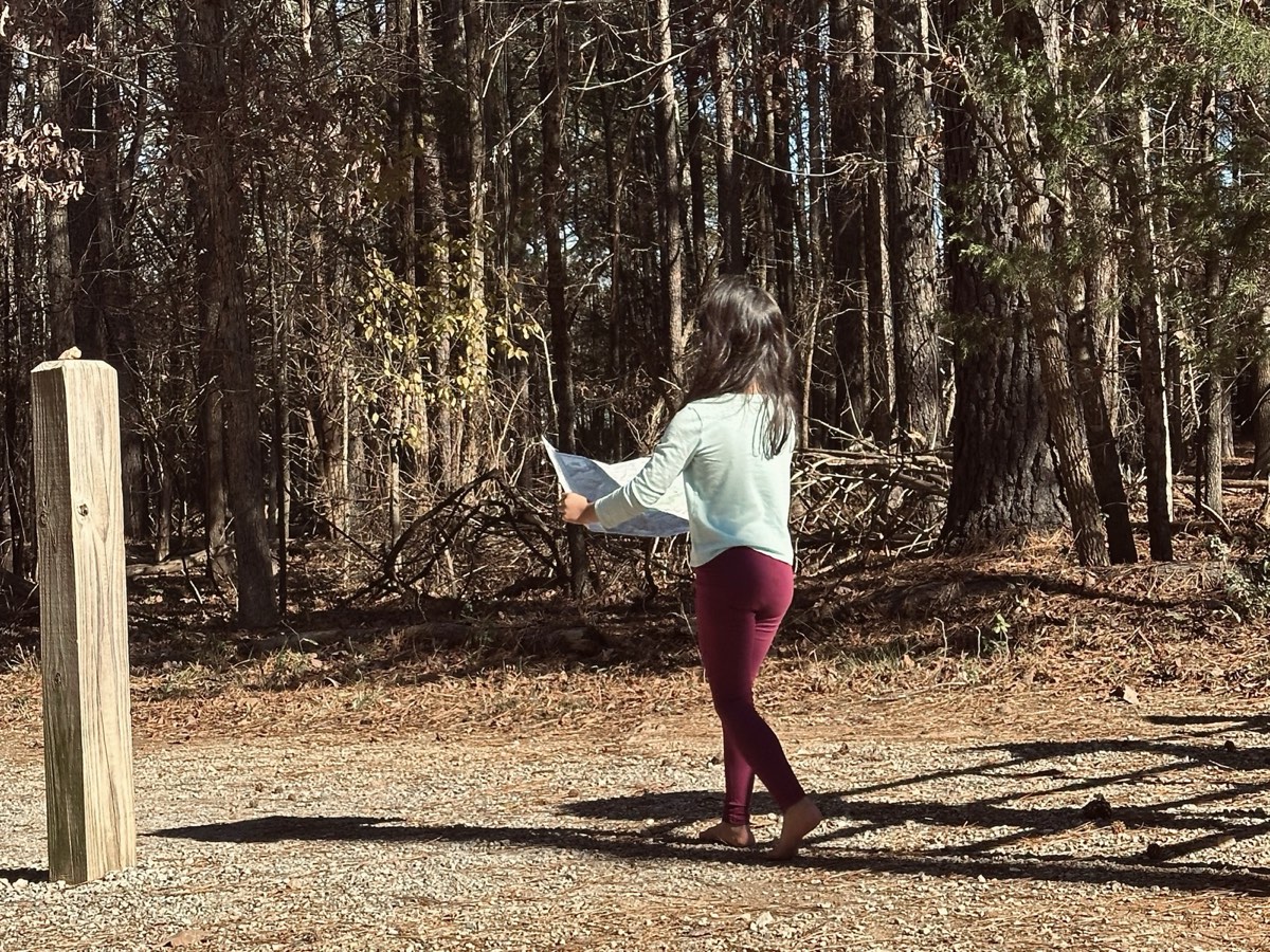 A child walking a gravel path at the edge of the woods, reading an open map